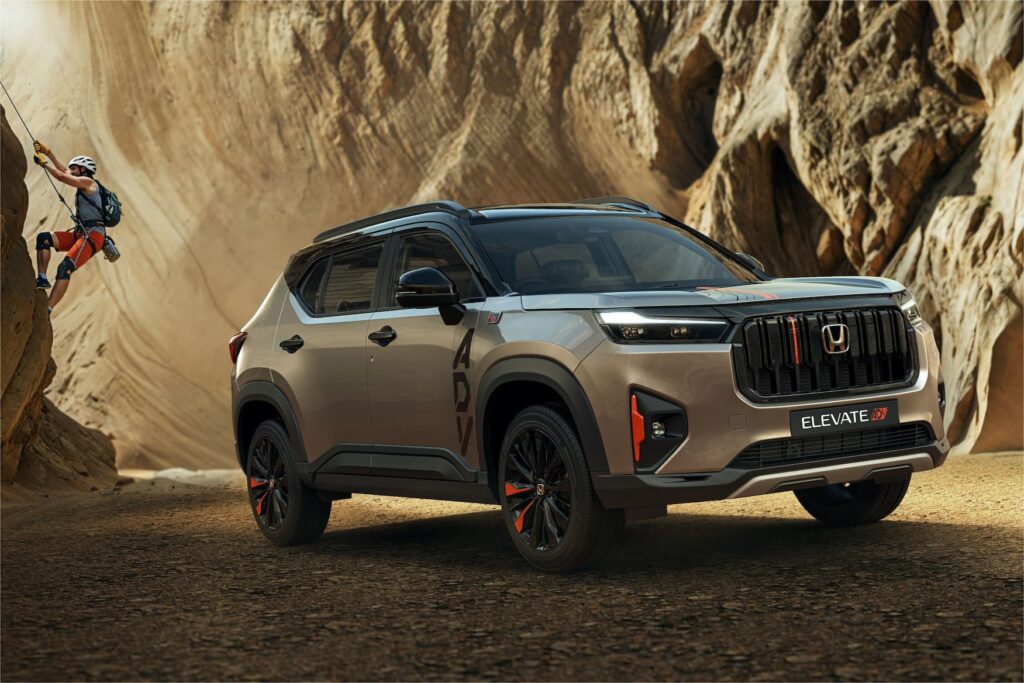 Front three-quarter view of the dual-tone Honda Elevate ADV (silver/grey and black) parked in a rugged, rocky canyon setting, featuring orange-accented wheels and a rock climber in the background.