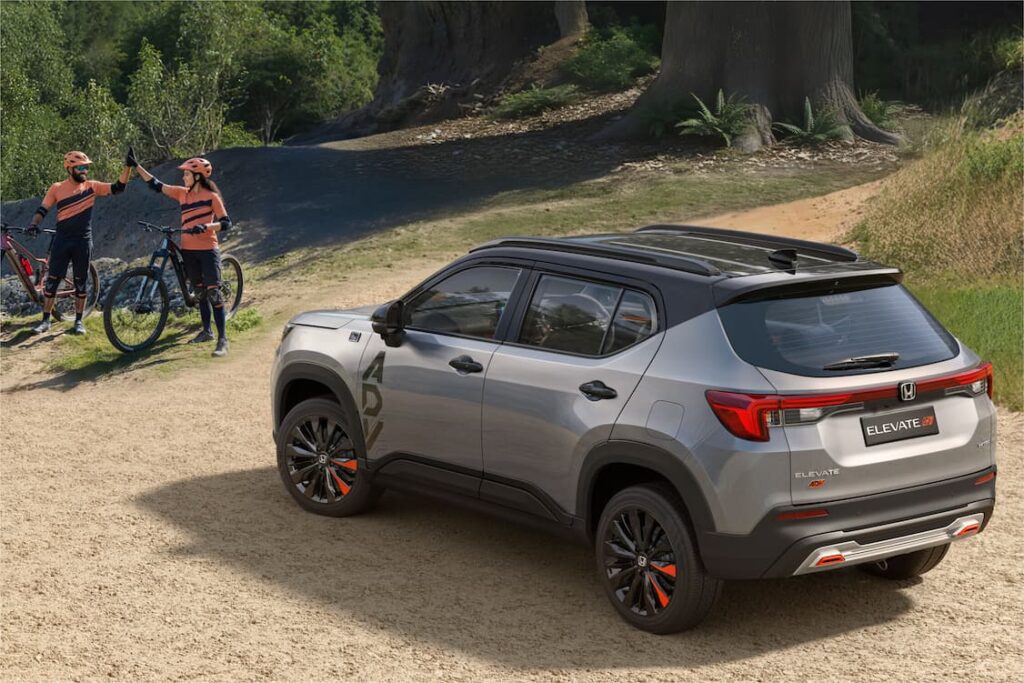 Rear three-quarter view of the dual-tone Honda Elevate ADV (silver/grey and black) on a dirt trail, featuring large 'ADV' side decals and black alloy wheels with orange accents. Two cyclists are standing nearby.